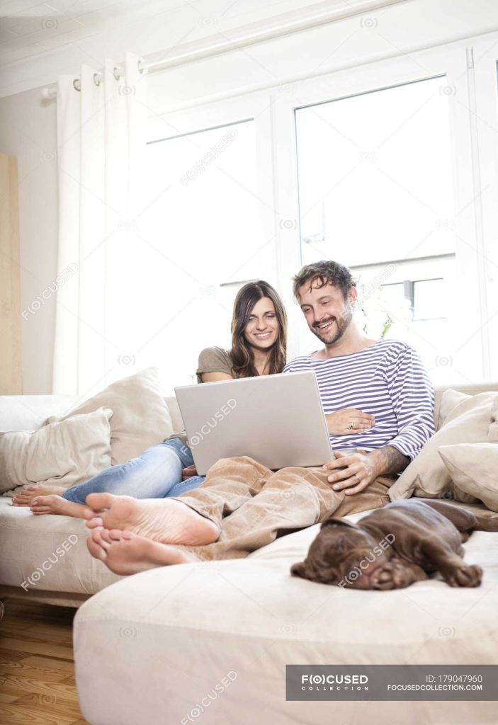 Улыбающаяся пара отдыхает с ноутбуком на диване в гостиной — человек, номер  - Stock Photo | #179047960